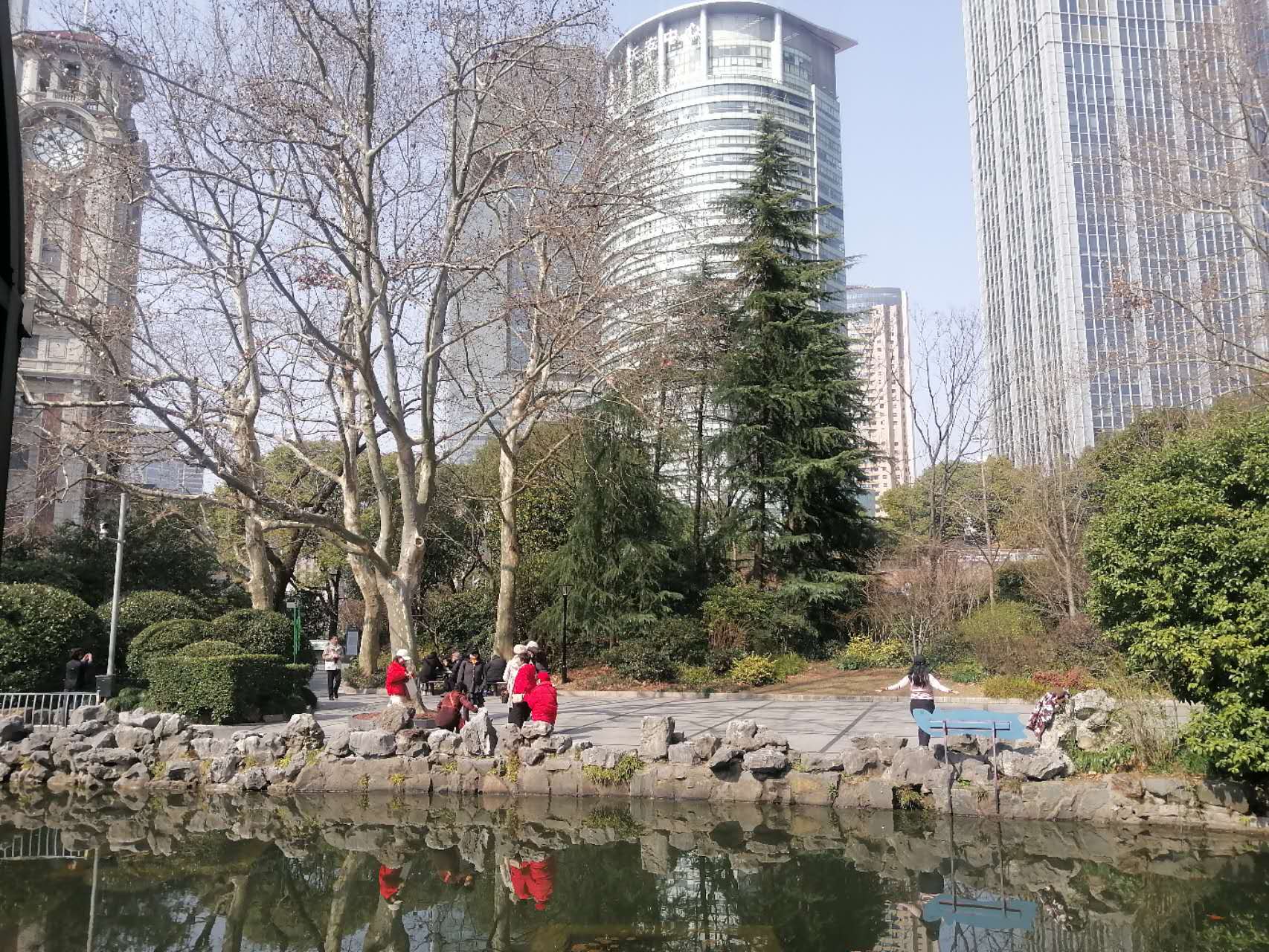 People beside a garden pond with tall city buildings behind the trees