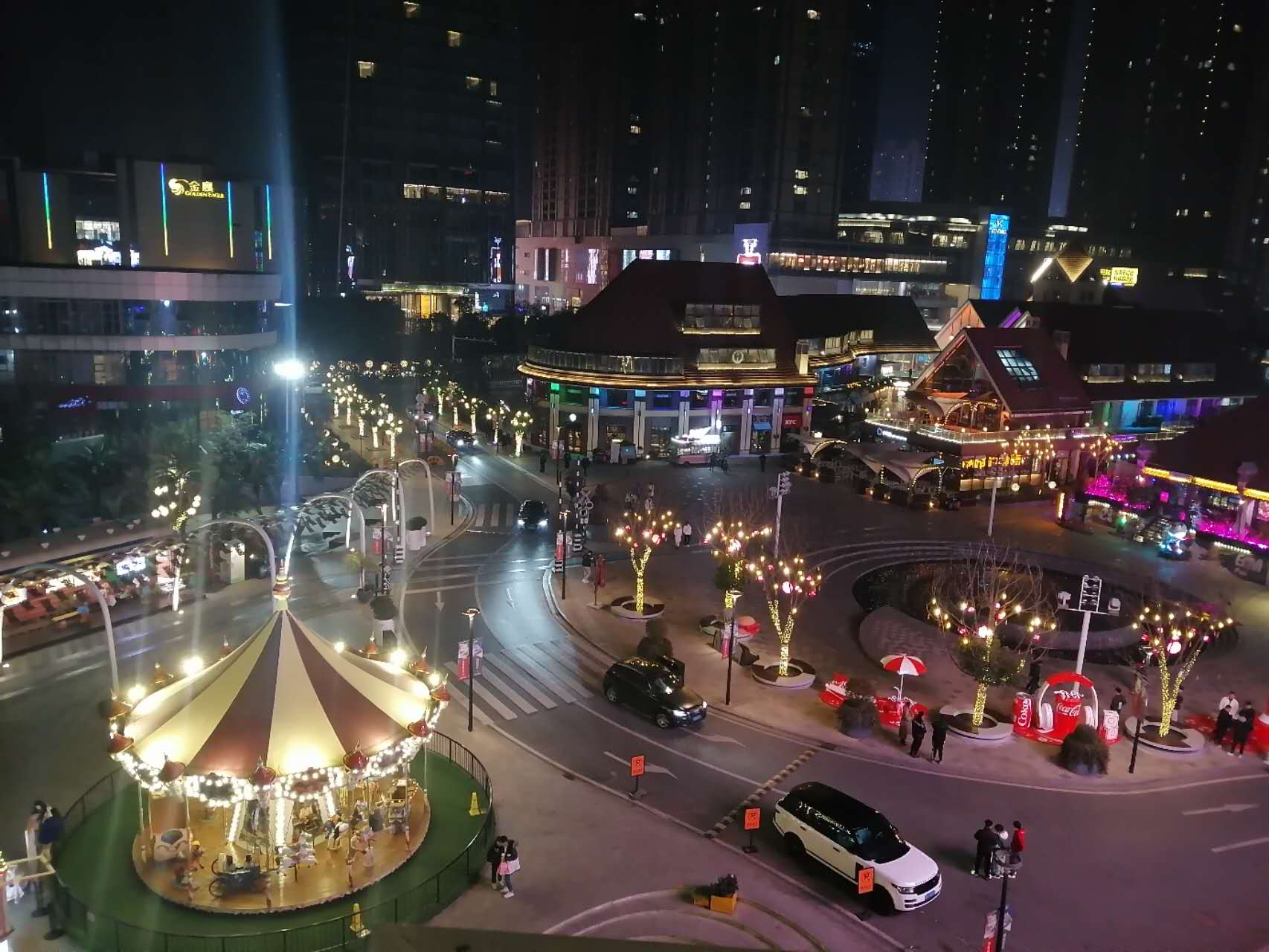 A brightly lit city square at night, with a carousel and restaurants